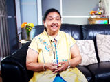 Mrs Parekh, smiling, sitting on her sofa while holding her lifeline pendant which is around her neck