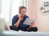 Lady sitting on herbed with her legs crossed, smiling holding her phone to her ear with her iPad in her other hand